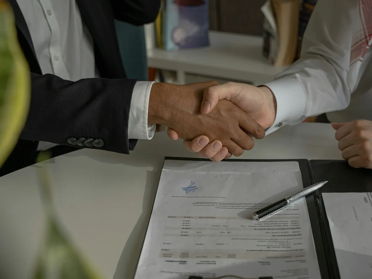 Two people shaking hands across a table, over a printed document and a pen