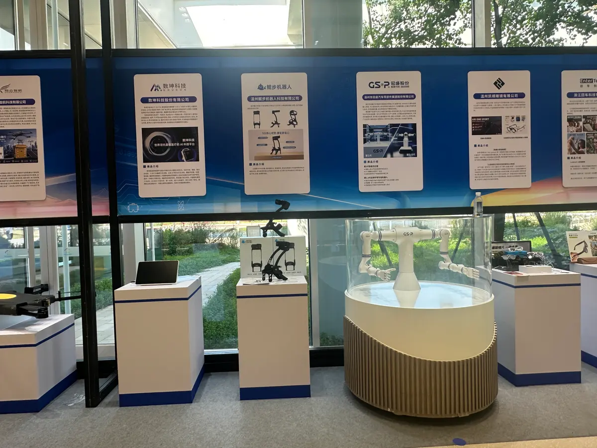 A white humanoid robotic arm in a round glass case, with an ankle exoskeleton, a tablet and AI glasses on neighbouring plinths